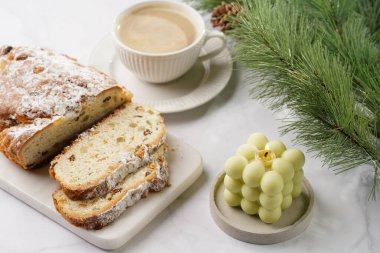 Beyaz tabak üzerinde kahve kapuçinosu olan beyaz porselen bardak, noel pastası çalıntısı ve mermer masada pastel yeşil baloncuk mumu.