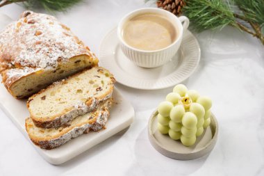 Beyaz tabak üzerinde kahve kapuçinosu olan beyaz porselen bardak, noel pastası çalıntısı ve mermer masada pastel yeşil baloncuk mumu.