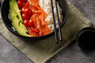 Poke kasesi - beyaz pirinçli siyah kase, tütsülenmiş somon, kiraz domatesleri ve avokado, susam tohumları, soya sosu ve gri beton bir yüzey üzerinde çubuklar, yakın plan.