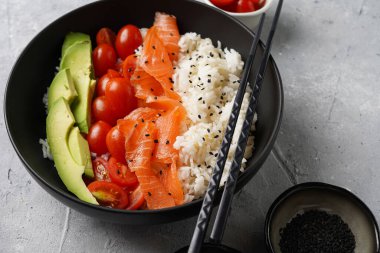 Poke kasesi - beyaz pirinçli siyah kase, tütsülenmiş somon, kiraz domatesleri ve avokado, susam tohumu, soya sosu ve gri beton yüzeyde çubuklar.