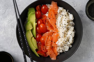Poke kasesi - beyaz pirinçli siyah kase, tütsülenmiş somon, kiraz domatesleri ve avokado, susam tohumları, soya sosu ve çubuklar gri beton yüzeyde, üst manzara