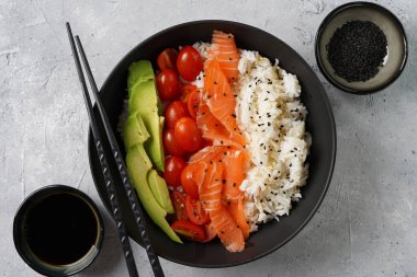 Poke kasesi - beyaz pirinçli siyah kase, tütsülenmiş somon, kiraz domatesleri ve avokado, susam tohumları, soya sosu ve çubuklar gri beton yüzeyde, üst manzara