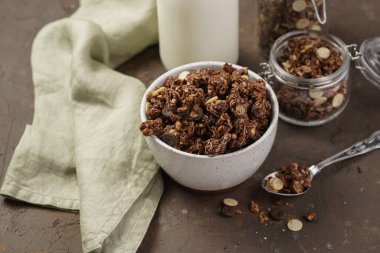 Kahvaltı müsli gevreği. Çikolata parçacıklı ve fındıklı. Birkaç kavanoz ve kâse, süt şişesi, kahverengi ahşap yüzey.