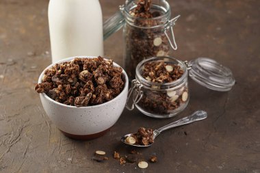 Kahvaltı müsli gevreği. Çikolata parçacıklı ve fındıklı. Birkaç kavanoz ve kâse, süt şişesi, kahverengi ahşap yüzey.