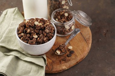 Kahvaltı müsli gevreği. Çikolata parçacıklı ve fındıklı. Birkaç kavanoz ve kâse, süt şişesi, kahverengi ahşap yüzey.