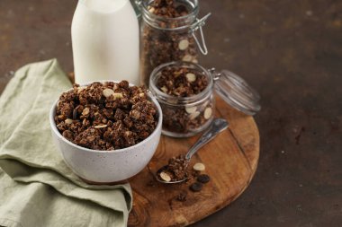 Kahvaltı müsli gevreği. Çikolata parçacıklı ve fındıklı. Birkaç kavanoz ve kâse, süt şişesi, kahverengi ahşap yüzey.