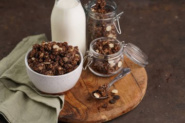 Kahvaltı müsli gevreği. Çikolata parçacıklı ve fındıklı. Birkaç kavanoz ve kâse, süt şişesi, kahverengi ahşap yüzey.