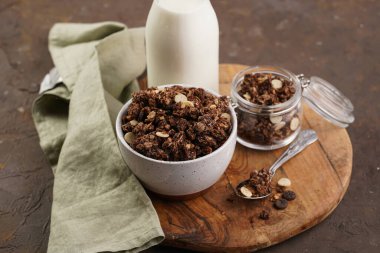 Kahvaltı müsli gevreği. Çikolata parçacıklı ve fındıklı. Birkaç kavanoz ve kâse, süt şişesi, kahverengi ahşap yüzey.