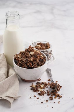 Kahvaltı müsli gevreği. Çikolata parçalı ve fındıklı. Birkaç kavanoz ve kâse, süt şişesi, beyaz yüzey.