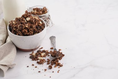 Kahvaltı müsli gevreği. Çikolata parçalı ve fındıklı. Birkaç kavanoz ve kâse, süt şişesi, beyaz yüzey.