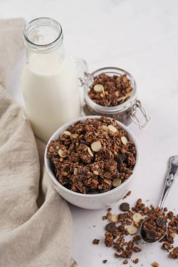 Kahvaltı müsli gevreği. Çikolata parçalı ve fındıklı. Birkaç kavanoz ve kâse, süt şişesi, beyaz yüzey.