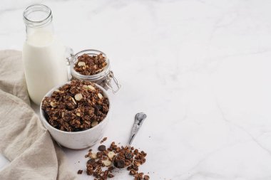 Kahvaltı müsli gevreği. Çikolata parçalı ve fındıklı. Birkaç kavanoz ve kâse, süt şişesi, beyaz yüzey.