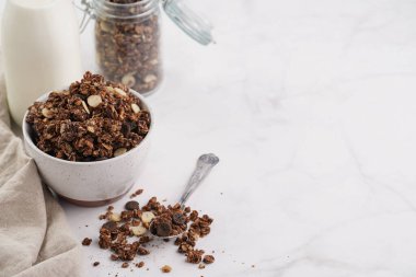 Kahvaltı müsli gevreği. Çikolata parçalı ve fındıklı. Birkaç kavanoz ve kâse, süt şişesi, beyaz yüzey.