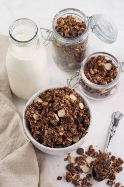 Kahvaltı müsli gevreği. Çikolata parçalı ve fındıklı. Birkaç kavanoz ve kâse, süt şişesi, beyaz yüzey.