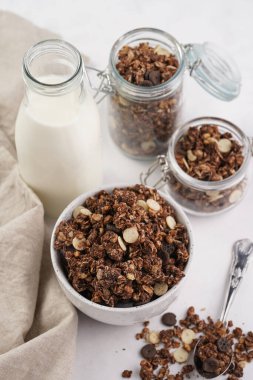 Kahvaltı müsli gevreği. Çikolata parçalı ve fındıklı. Birkaç kavanoz ve kâse, süt şişesi, beyaz yüzey.