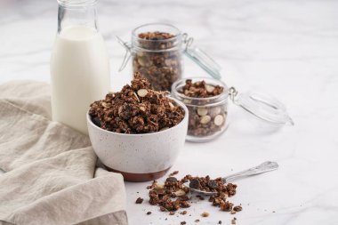 Kahvaltı müsli gevreği. Çikolata parçalı ve fındıklı. Birkaç kavanoz ve kâse, süt şişesi, beyaz yüzey.