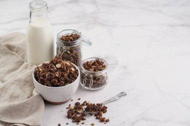 Kahvaltı müsli gevreği. Çikolata parçalı ve fındıklı. Birkaç kavanoz ve kâse, süt şişesi, beyaz yüzey.