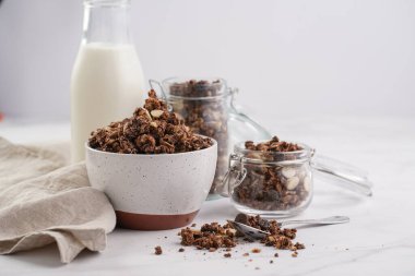 Kahvaltı müsli gevreği. Çikolata parçalı ve fındıklı. Birkaç kavanoz ve kâse, süt şişesi, beyaz yüzey.