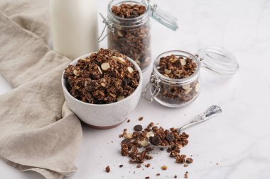 Kahvaltı müsli gevreği. Çikolata parçalı ve fındıklı. Birkaç kavanoz ve kâse, süt şişesi, beyaz yüzey.