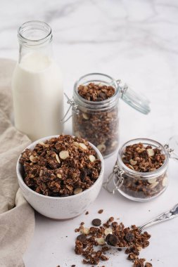 Kahvaltı müsli gevreği. Çikolata parçalı ve fındıklı. Birkaç kavanoz ve kâse, süt şişesi, beyaz yüzey.