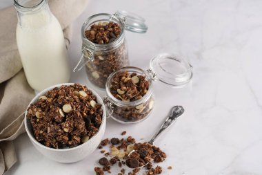 Kahvaltı müsli gevreği. Çikolata parçalı ve fındıklı. Birkaç kavanoz ve kâse, süt şişesi, beyaz yüzey.