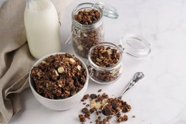 Kahvaltı müsli gevreği. Çikolata parçalı ve fındıklı. Birkaç kavanoz ve kâse, süt şişesi, beyaz yüzey.