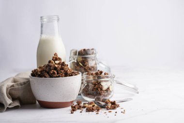 Kahvaltı müsli gevreği. Çikolata parçalı ve fındıklı. Birkaç kavanoz ve kâse, süt şişesi, beyaz yüzey.