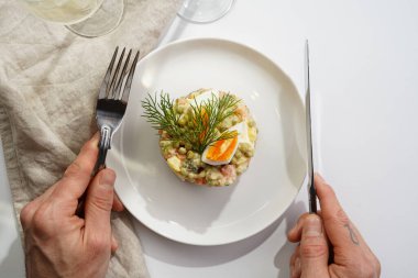 Yeni yıl arifesi için geleneksel Doğu Avrupa tabağının üzerinde gümüş çatal ve bıçak tutan erkek el - patates, havuç, soğan, bezelye, sosis, salatalık, yumurta ve mayonez salatası beyaz bir tabakta 