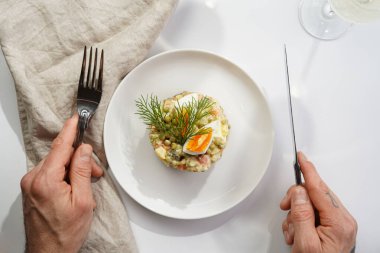 Yeni yıl arifesi için geleneksel Doğu Avrupa tabağının üzerinde gümüş çatal ve bıçak tutan erkek el - patates, havuç, soğan, bezelye, sosis, salatalık, yumurta ve mayonez salatası beyaz bir tabakta 