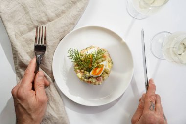 Yeni yıl arifesi için geleneksel Doğu Avrupa tabağının üzerinde gümüş çatal ve bıçak tutan erkek el - patates, havuç, soğan, bezelye, sosis, salatalık, yumurta ve mayonez salatası beyaz bir tabakta 