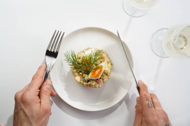 Yeni yıl arifesi için geleneksel Doğu Avrupa tabağının üzerinde gümüş çatal ve bıçak tutan erkek el - patates, havuç, soğan, bezelye, sosis, salatalık, yumurta ve mayonez salatası beyaz bir tabakta 