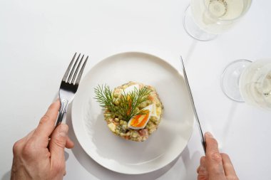 Yeni yıl arifesi için geleneksel Doğu Avrupa tabağının üzerinde gümüş çatal ve bıçak tutan erkek el - patates, havuç, soğan, bezelye, sosis, salatalık, yumurta ve mayonez salatası beyaz bir tabakta 