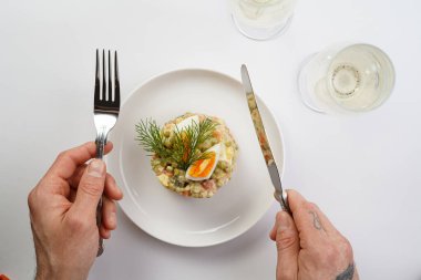 Yeni yıl arifesi için geleneksel Doğu Avrupa tabağının üzerinde gümüş çatal ve bıçak tutan erkek el - patates, havuç, soğan, bezelye, sosis, salatalık, yumurta ve mayonez salatası beyaz bir tabakta 