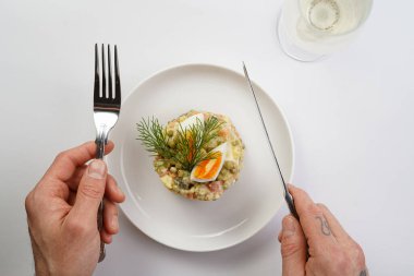 Yeni yıl arifesi için geleneksel Doğu Avrupa tabağının üzerinde gümüş çatal ve bıçak tutan erkek el - patates, havuç, soğan, bezelye, sosis, salatalık, yumurta ve mayonez salatası beyaz bir tabakta 