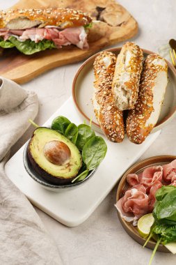 Lezzetli kahvaltı malzemeleri: yulaf ve tohumla süslenmiş kahverengi küllü ekmek çubukları, domuz dilimleri prosciutto, sert peynirli gouda ve mermer tahtada taze ıspanak ve avokado.