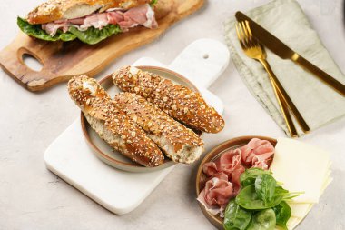 Lezzetli kahvaltı malzemeleri: yulaf ve tohumlarla süslenmiş kahverengi küllü ekmek çubukları, domuz dilimleri prosciutto, sert peynirli gouda ve mermer tahtada taze ıspanak yaprakları