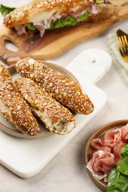 Lezzetli kahvaltı malzemeleri: yulaf ve tohumla süslenmiş kahverengi küllü ekmek çubukları, domuz dilimleri prosciutto, mermer tahtada sert peynir gouda
