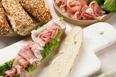 Yulaf ezmeli, domuz dilimli prosciutto, sert peynirli gouda ve mermer tahtada taze yeşillik, bej yüzeyle süslenmiş kahverengi küllü ekmek çubuklarıyla uzun sandviç.