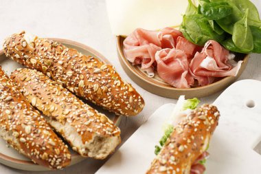 Yulaf ezmeli, domuz dilimli prosciutto, sert peynirli gouda ve mermer tahtada taze yeşillik, bej yüzeyle süslenmiş kahverengi küllü ekmek çubuklarıyla uzun sandviç.