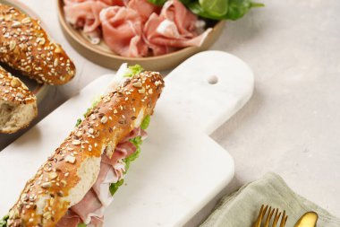 Yulaf ezmeli, domuz dilimli prosciutto, sert peynirli gouda ve mermer tahtada taze yeşillik, bej yüzeyle süslenmiş kahverengi küllü ekmek çubuklarıyla uzun sandviç.