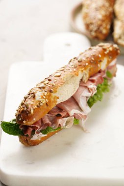 Yulaf ezmeli, domuz dilimli prosciutto, sert peynirli gouda ve mermer tahtada taze yeşillik, bej yüzeyle süslenmiş kahverengi küllü ekmek çubuklarıyla uzun sandviç.