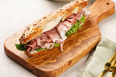 Kahverengi küllü ekmek çubuklarıyla süslenmiş uzun sandviç yulaf, domuz dilimleri prosciutto, sert peynirli gouda ve ahşap tahtada taze yeşiller, bej yüzey.