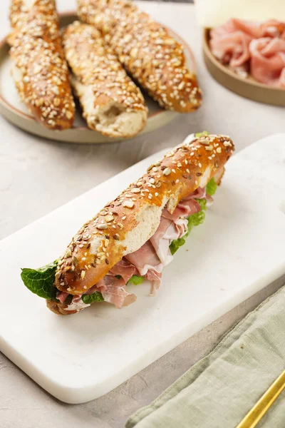 Long sandwich in brown lye bread sticks garnished with oats , pork slices prosciutto, hard cheese gouda and fresh greens on marble board, beige surface