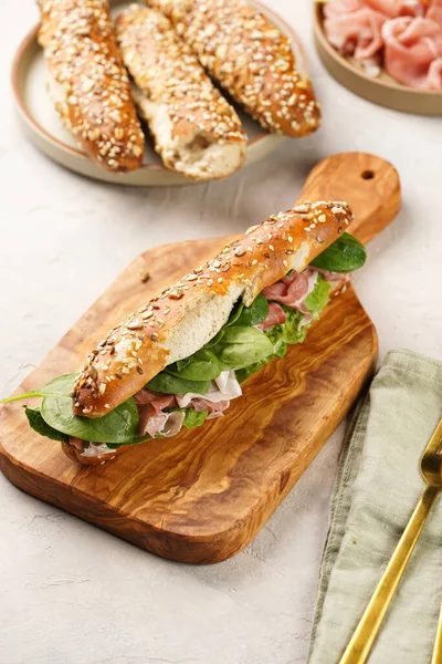 Long sandwich in brown lye bread sticks garnished with oats , pork slices prosciutto, hard cheese gouda and fresh greens on wooden board, beige surface