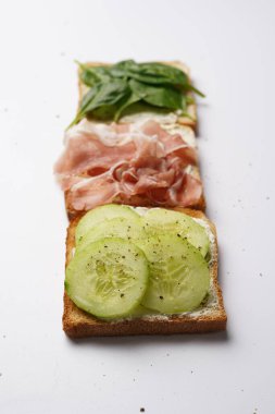 Beyaz bir yüzeyde krem peynirli, jambonlu, salatalıklı ve ıspanaklı kızarmış ekmek.