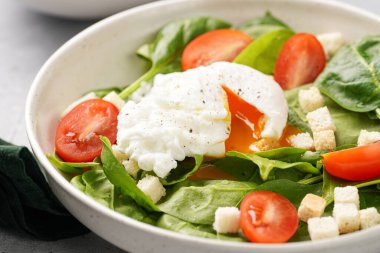 Taze yeşil ıspanaklı salata kasesi, kiraz domatesleri, kıtır ekmekler, yumurtalı krema sosu ve gri taşlı bir yüzey.