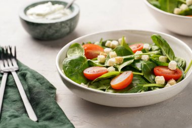 Taze yeşil ıspanaklı salata kasesi, kiraz domatesleri, kızarmış ekşi krema sosu ve gri taşlı bir yüzey.