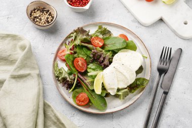 Yeşil yapraklı salata, marul, ıspanak, kiraz domatesleri, mozarella, zeytinyağı, biber, gri taş yüzeyde limon suyu.