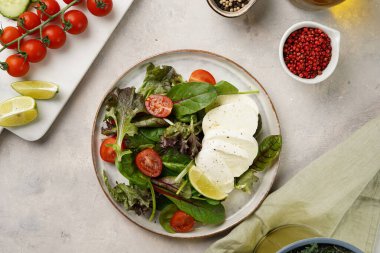Yeşil yapraklı salata, marul, ıspanak, kiraz domatesleri, mozarella, zeytinyağı, kırmızı biber, gri taş yüzeyde limon suyu, üst manzara.