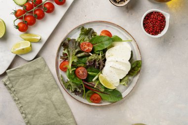 Yeşil yapraklı salata, marul, ıspanak, kiraz domatesleri, mozarella, zeytinyağı, kırmızı biber, gri taş yüzeyde limon suyu, üst manzara.